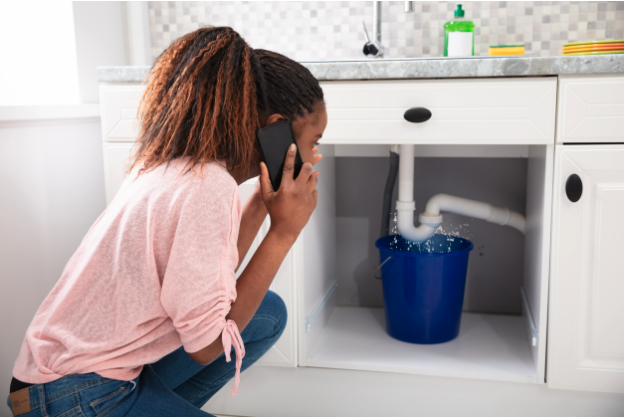 Woman calling plumbing in front of leaking kitchen sink pipe.